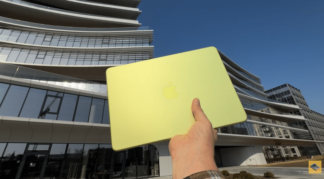 MacBook Neo лимонного кольору в руці на фоні сучасної будівлі