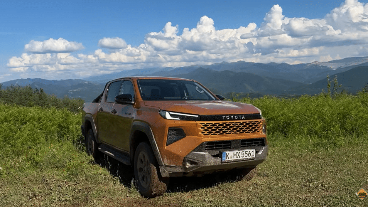 Toyota Hilux 2026 в помаранчевому кольорі на гірському бездоріжжі на тлі зелених схилів і блакитного неба