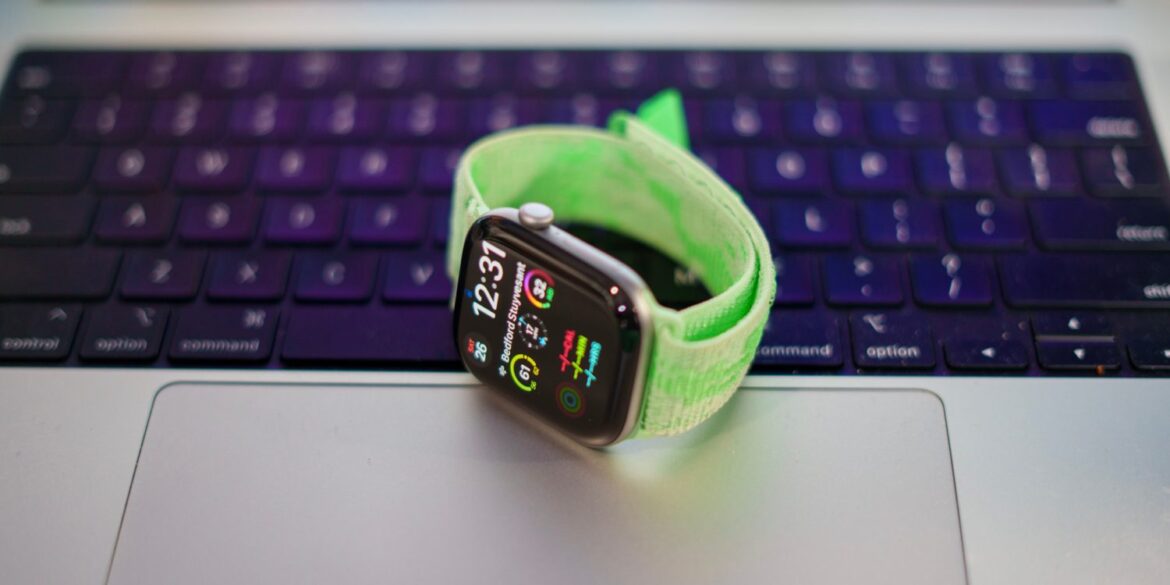 Apple Watch Series 12 з зеленим ремінцем на клавіатурі MacBook, екран відображає час 1:23 та різноманітні віджети