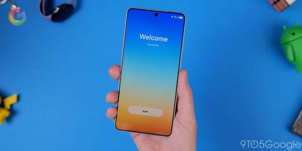 Смартфон в руці з екраном приветствія One UI 9 на синьому фоні з навколишніми гаджетами