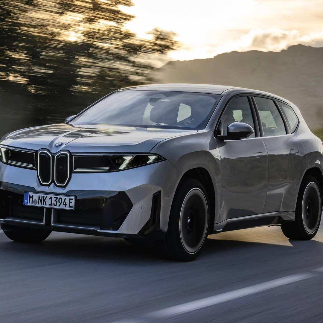Сріблястий BMW iX3 їде по дорозі на тлі гір у сонячну погоду