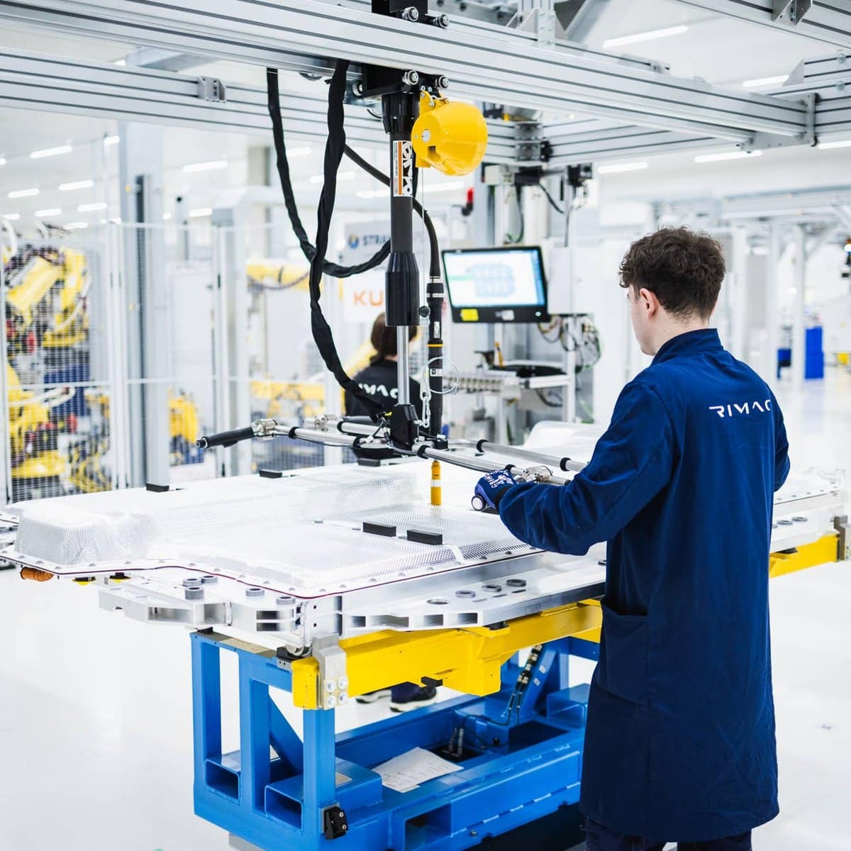 Працівник у синьому робочому комбінезоні з логотипом Rimac обробляє батарейний модуль на складальному стенді у світлому цеху