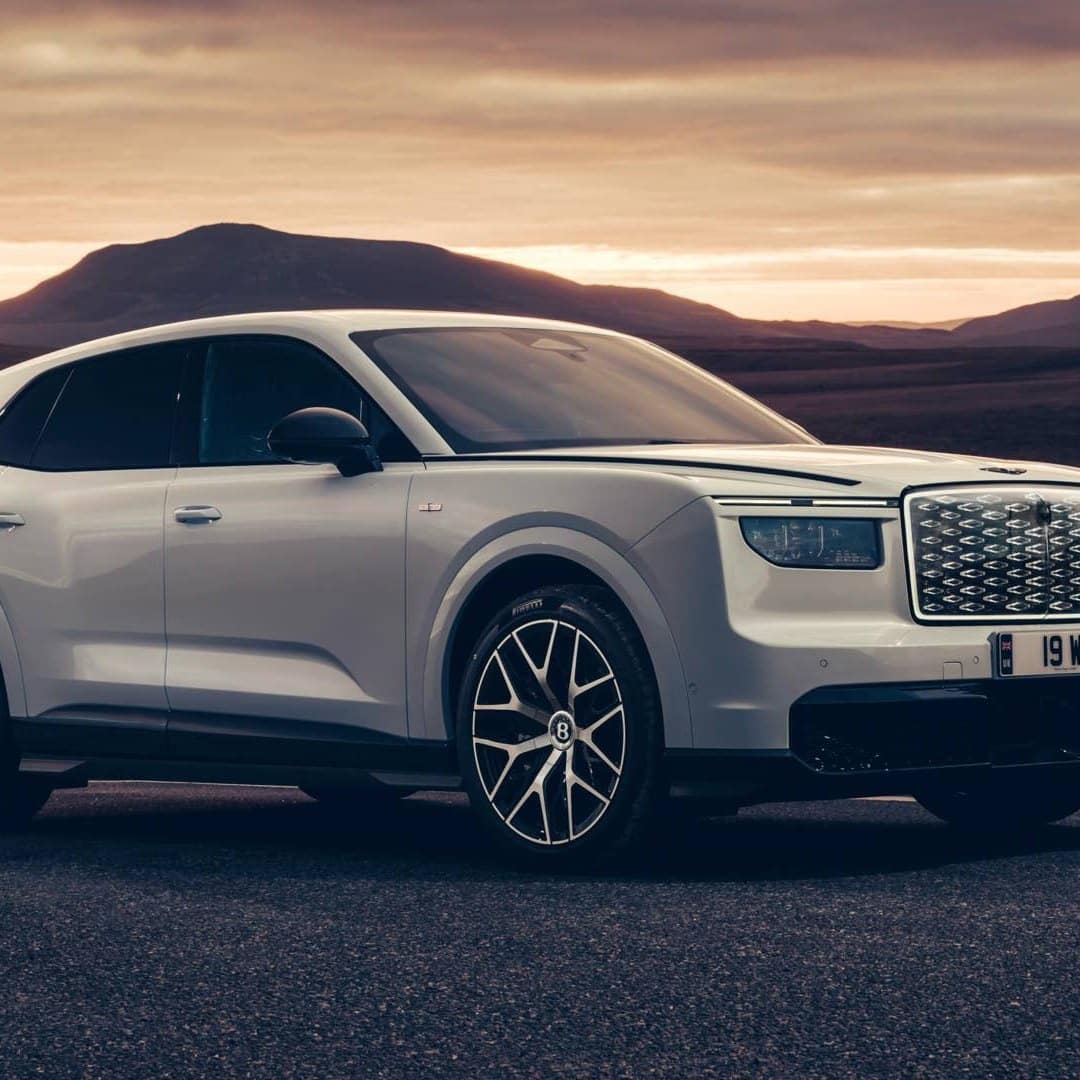 Білий Bentley Torcal у русі на тлі заходу сонця біля дороги серед пустельних пагорбів із ромбовою решіткою радіатора