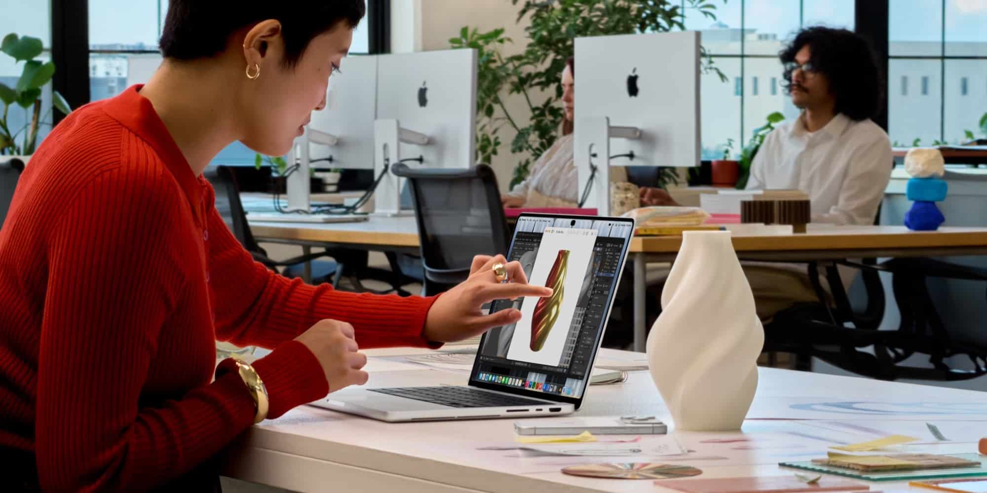 Жінка в червоному светрі торкається пальцем екрана MacBook Pro з 3D-моделлю у світлому офісі