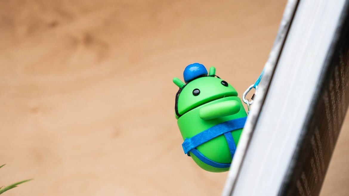 Зелена фігурка робота Android із синім ковпачком і синім ременем на тілі, прикріплена карабіном до білої поверхні