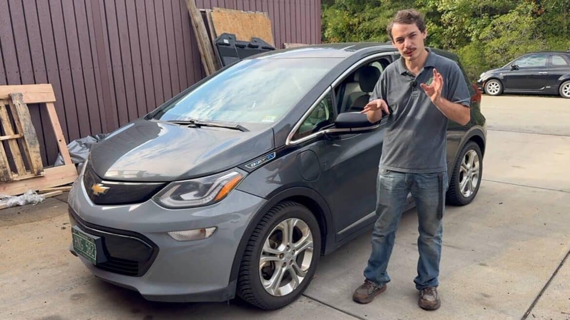 Чоловік у сірій сорочці жестикулює біля сірого Chevrolet Bolt EV на подвір'ї