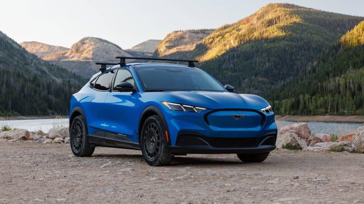 Синій електрокросовер Ford Mustang Mach-E Trail з чорними колесами стоїть на гравійному березі озера на тлі гір і хвойних