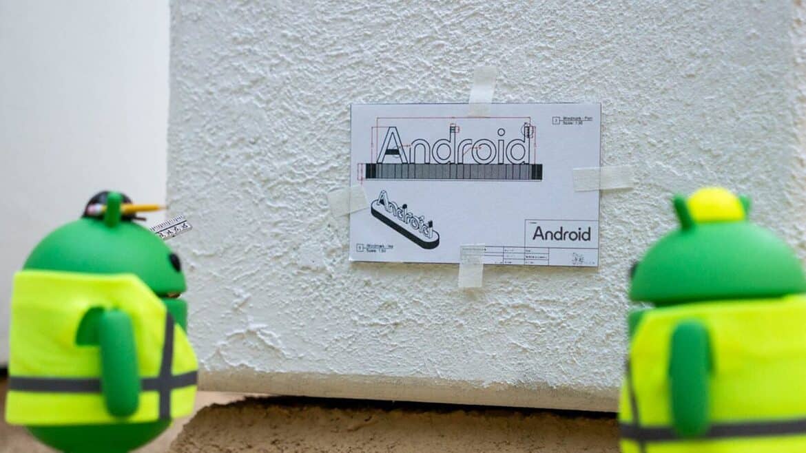 Зелені фігурки Android у жовтих жилетах біля білої стіни з прикріпленим кресленням логотипу
