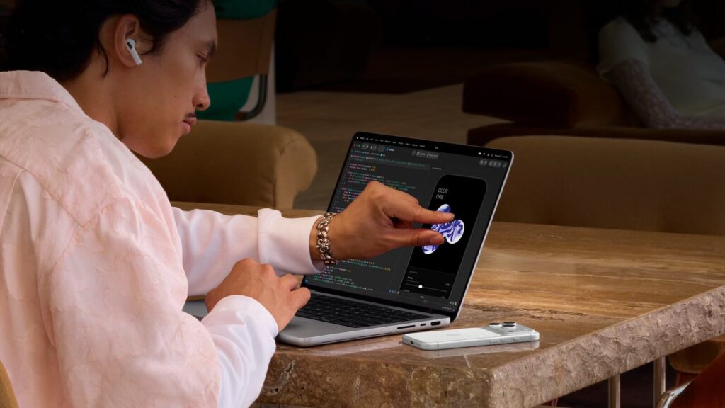 Чоловік у рожевій сорочці з навушником торкається екрана MacBook Pro з кодом на дерев'яному столі